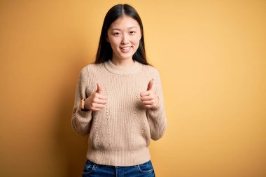 Sarı izole edilmiş arka plan başarısı işareti üzerine sıradan süveter giyen genç, güzel Asyalı bir kadın. Elleriyle olumlu jest yapıyor. Gülümsüyor ve mutlu. Neşeli ifade ve kazanan jest.