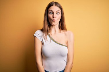 İzole edilmiş sarı arka plan üzerine gündelik tişört giyen genç, güzel, kızıl saçlı kadın dudaklı balık suratı yapıyor, çılgınca ve komik bir jest. Komik ifade.