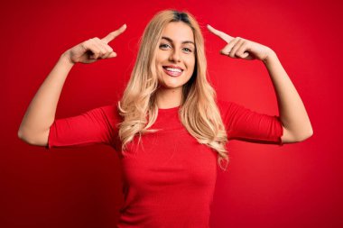 Genç, güzel, sarışın bir kadın, gündelik tişört giyiyor, kırmızı arka planda dikiliyor, gülümsüyor, iki eliyle başını işaret ediyor, harika bir fikir ya da düşünce, iyi bir hafıza.