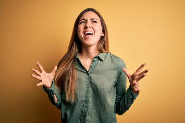 Sarı arka planda yeşil tişörtlü, sarışın, genç, güzel bir kadın. Deli deli bağırıyor, agresif bir ifadeyle bağırıyor ve kollarını kaldırıyor. Hayal kırıklığı konsepti.