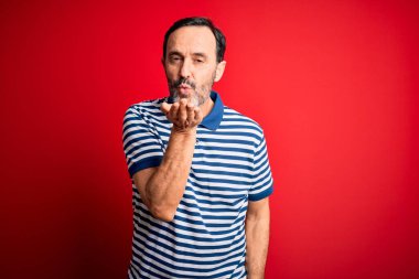 Orta yaşlı, beyaz, sıradan çizgili polo giyen, izole edilmiş kırmızı arka planda dikilip havada öpücükler saçan, güzel ve seksi bir adam. Aşk ifadesi.