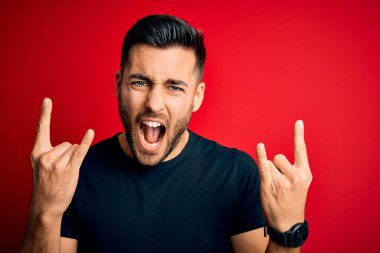 Siyah bir tişört giyen yakışıklı genç adam izole edilmiş kırmızı arka planda duruyor çılgın bir ifadeyle elleri havada rock sembolü yapıyor diye bağırıyor. Müzik yıldızı. Ağır konsept.