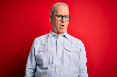 Orta yaşlı yakışıklı, beyaz bir adam kırmızı arka planda sıradan çizgili gömlek ve gözlük takıyor. Şok olmuş, şüpheci ve alaycı görünüyor, ağzı açık şaşırmış.
