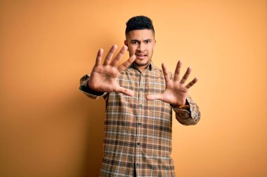 Izole sarı arka plan üzerinde duran rahat gömlek giyen genç yakışıklı adam korkmuş ve korku ifade ile korkmuş elleriyle jest durdurmak, şok içinde bağırarak. Panik kavramı.