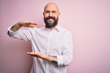 Sakallı, sakallı, zarif tişörtlü, soyutlanmış pembe arka planda, elleri büyük ve büyük boy işareti gösteren, ölçü sembolü olan yakışıklı kel bir adam. Kameraya bakıp gülümsüyor. Ölçüm konsepti.