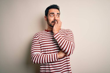 Sakallı, sıradan çizgili tişörtlü, beyaz arka planda duran, gergin ve gergin, ağzı tırnak yiyen genç yakışıklı bir adam. Endişe sorunu.