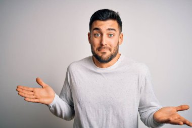 İzole edilmiş beyaz arka plan üzerinde duran sıradan bir tişört giyen genç yakışıklı ve elleri ve kolları havada, kafası karışmış bir ifade. Şüphe kavramı.