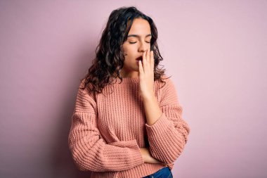 Kıvırcık saçlı, izole edilmiş pembe arka planda günlük süveter giyen genç ve güzel bir kadın ağzıyla esnemekten sıkılmış. Huzursuz ve uykulu..