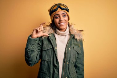 Genç Afrikalı Afro-Amerikan kayakçı kız kar sporu kıyafetleri ve kayak gözlüğü takıyor gülümsüyor ve el hareketleriyle el hareketi yapıyor parmakları ve kamerasıyla küçük boy işareti yapıyor. Ölçü kavramı.