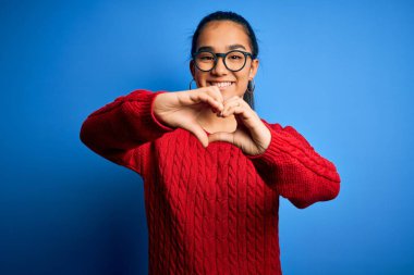 Genç, güzel Asyalı bir kadın mavi arka planda gözlüklü, sıradan bir süveter giyiyor. Aşkla gülümsüyor, elleriyle kalp sembolü yapıyor. Romantik konsept.