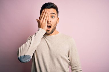 Genç, yakışıklı bir adam, günlük süveter giyiyor, pembe arka planda duruyor, bir gözünü eliyle kapatıyor, yüzünde kendine güvenen bir gülümseme ve sürpriz bir duygu var..