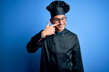 Genç, yakışıklı aşçı üniforması ve izole edilmiş mavi arka plan üzerinde şapkası olan bir adam. El parmağı ve burunla işaret ediyor. Neşeli gülümsüyor. Güzellik konsepti