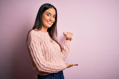 Genç, güzel, esmer bir kadın pembe arka plan üzerine günlük süveter giyip gülümseyerek içeri girmeye davet ediyor.