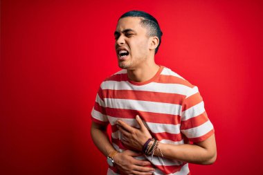 Hafif çizgili tişört giyen genç Brezilyalı adam midesi bulandığı için el üstünde kırmızı arka plan üzerinde dikiliyor. Ache kavramı.
