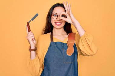 Önlük takan genç, güzel bir berber kadın. Sarı arka planda jilet taşıyor. Gülümsüyor.