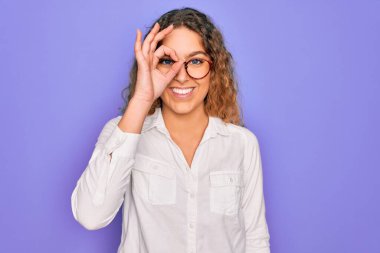 Mavi gözlü genç bir kadın mor arka planda günlük gömlek ve gözlük takıyor. El hareketleriyle gülümsüyor ve mutlu yüzle parmaklarının arasından bakıyor..