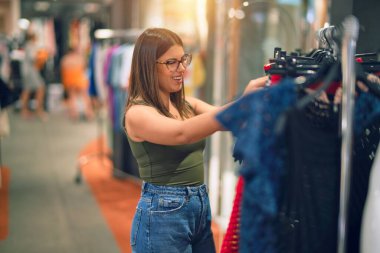 Young beautiful woman smiling happy and confident. Standing with smile on face looking clothes at store