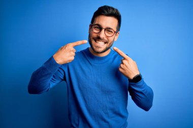 Sakallı, sıradan süveter giyen, mavi arka planda gözlüklü neşeli gülümseyen ve parmaklarıyla ve ağzıyla işaret eden genç yakışıklı bir adam. Diş sağlığı konsepti.