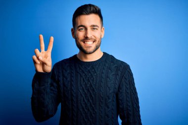 İzole edilmiş mavi arka plan üzerinde duran sıradan süveter giyen genç ve yakışıklı bir adam iki numaralı parmaklarıyla işaret ederken kendinden emin ve mutlu bir şekilde gülümsüyor..