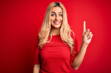 Genç ve güzel sarışın bir kadın, günlük tişört giyip kırmızı arka planda dikiliyor. Bir numaralı parmağını gösterip gülümsüyor ve mutlu oluyor..