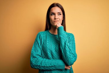 Sarı arka planda yeşil süveter giyen genç, güzel esmer kadın. Elleri çenesinde, soru sormayı düşünüyor. Düşünceli bir ifade. Düşünceli bir yüzle gülümsüyor. Şüphe kavramı.