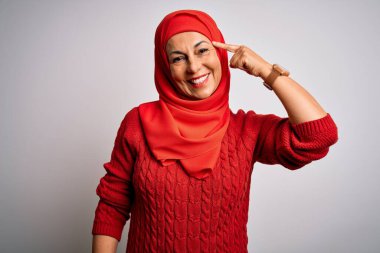 Orta yaşlı esmer kadın izole edilmiş beyaz arka plan üzerine geleneksel başörtüsü takıyor. Tek parmakla başını göstererek gülümsüyor. Harika bir fikir ya da düşünce, iyi bir hafıza.