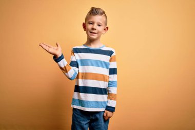 Küçük, beyaz, mavi gözlü, sarı arka planda renkli çizgili gömlek giyen neşeli bir şekilde gülümseyen, eli kameraya bakan bir çocuk..