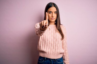 Genç, güzel, esmer bir kadın pembe arka plan üzerine günlük süveter giyiyor. Parmağını kaldırıyor ve kızgın bir ifade sergiliyor.
