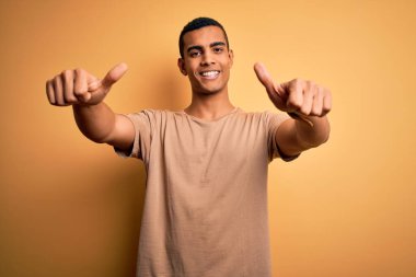 Sarı arka plan üzerinde sıradan tişörtler giyen genç ve yakışıklı afro-amerikan adam el hareketleriyle olumlu jest yapıyor, gülümsüyor ve başarı için mutlu. Kazanan jest.