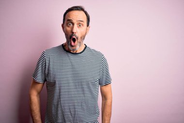 Orta yaşlı, beyaz, sıradan çizgili tişört giyen, pembe arka planda duran, korku, korku ve heyecan dolu bir yüz ifadesinden korkan ve şok olan bir adam..