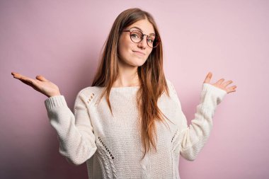 Genç, güzel, kızıl saçlı bir kadın pembe arka planda gözlük ve süveter takıyor. Kolları ve elleri havada, kafası karışmış bir ifade takınıyor. Şüphe kavramı.