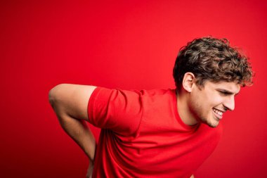 Sarışın, kıvırcık saçlı, kırmızı arka planda sıradan tişörtler giyen, sırt ağrısı çeken, elleriyle dokunan, kaslı bir erkek.