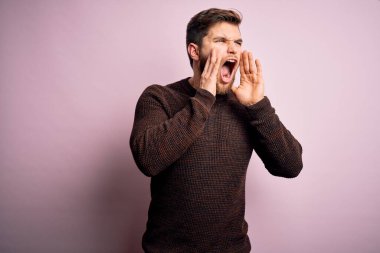 Sakallı ve mavi gözlü genç bir adam pembe arka planda sıradan bir süveter giyiyor ve elleri ağzında sinirli bir şekilde bağırıyor.