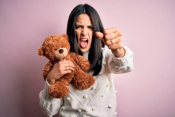 Young brunette woman with blue eyes hugging teddy bear stuffed animal over pink background annoyed and frustrated shouting with anger, crazy and yelling with raised hand, anger concept