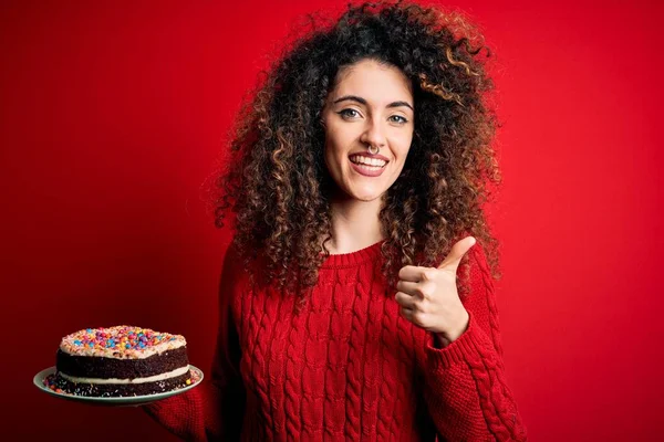 Young beautiful woman with curly hair and piercing holding plate with birthday cake happy with big smile doing ok sign, thumb up with fingers, excellent sign