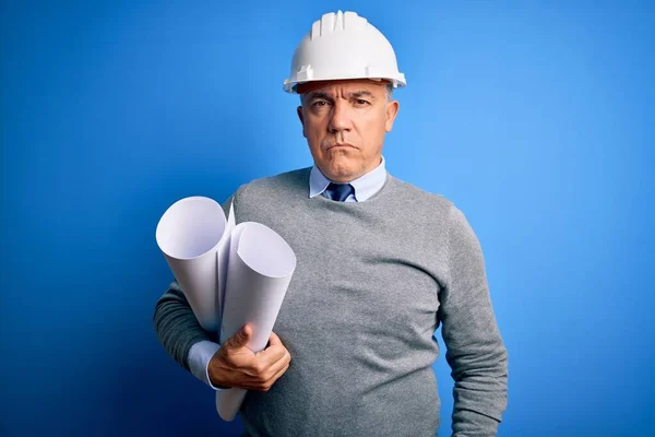Middle age handsome grey-haired architect man wearing safety helmet holding blueprints Relaxed with serious expression on face. Simple and natural looking at the camera.