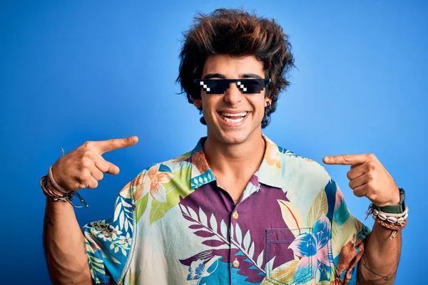 Young handsome man wearing thug life glasses standing over isolated blue background looking confident with smile on face, pointing oneself with fingers proud and happy.