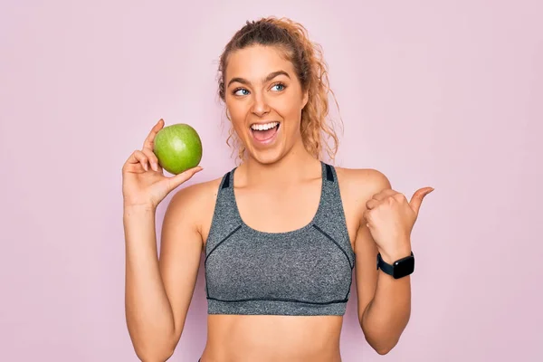 年轻美丽的金发女运动员 蓝眼睛 手握健康的绿色苹果果实 手握大拇指 笑容满面地站在一旁 — 图库照片
