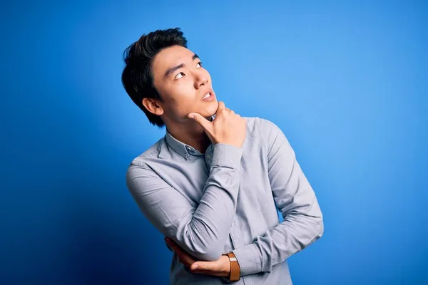 年轻英俊的中国男人穿着休闲装 站在孤立的蓝色背景下思考着一个问题 手托着下巴忧心忡忡 — 图库照片
