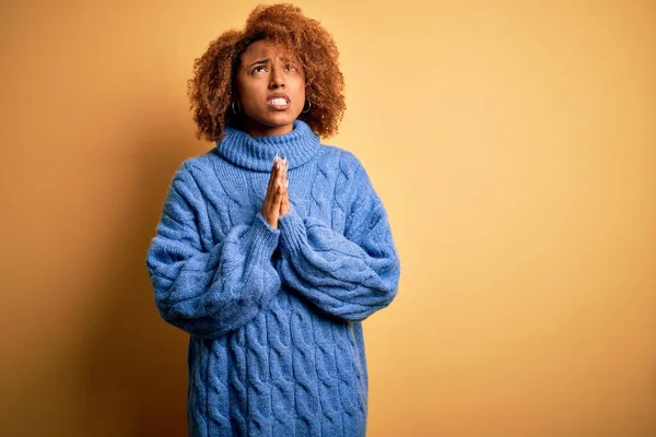 Young beautiful African American afro woman with curly hair wearing blue turtleneck sweater begging and praying with hands together with hope expression on face very emotional and worried. Begging.