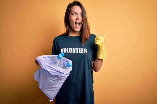 Beautiful volunteer girl caring environment doing volunteering holding bag with rubish bottles pointing and showing with thumb up to the side with happy face smiling