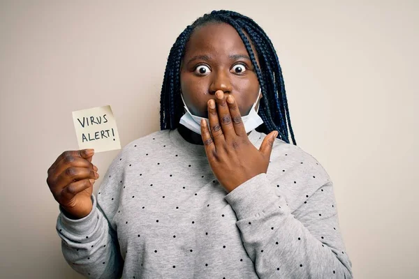 African american woman wearing medical mask holding reminder with virus alert message cover mouth with hand shocked with shame for mistake, expression of fear, scared in silence, secret concept