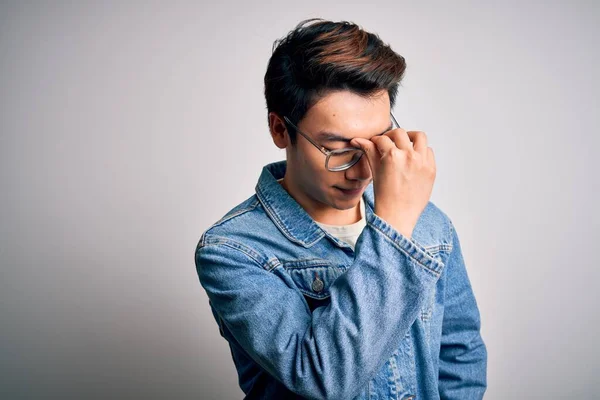 Young handsome chinese man wearing denim jacket and glasses over white background tired rubbing nose and eyes feeling fatigue and headache. Stress and frustration concept.