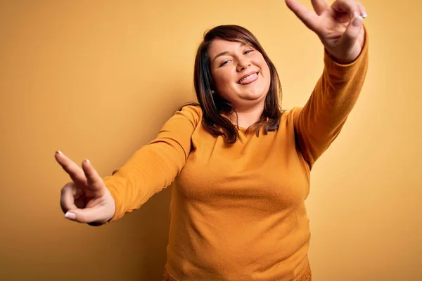 Beautiful brunette plus size woman wearing casual sweater over isolated yellow background smiling with tongue out showing fingers of both hands doing victory sign. Number two.