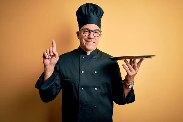 Middle age handsome grey-haired waiter man wearing cooker uniform and hat holding tray surprised with an idea or question pointing finger with happy face, number one