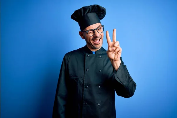 年轻英俊的厨师 身穿炊事服 头戴一顶帽子 头戴孤立的蓝色背景 喜形于色地微笑着 用手指在镜头前眨眼示意胜利 第二点 — 图库照片