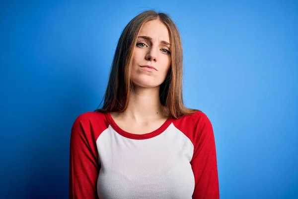 隔離された青の背景にカジュアルなTシャツを着た若い美しい赤毛の女性が顔に深刻な表情でリラックスしています シンプルで自然なカメラを見て — ストック写真