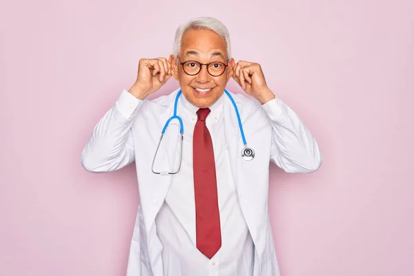 中世のシニア白髪の医者の男性は 聴診器とプロの医療用コートを身に着けています指で耳を引く笑顔 面白いジェスチャー オーディション問題 — ストック写真
