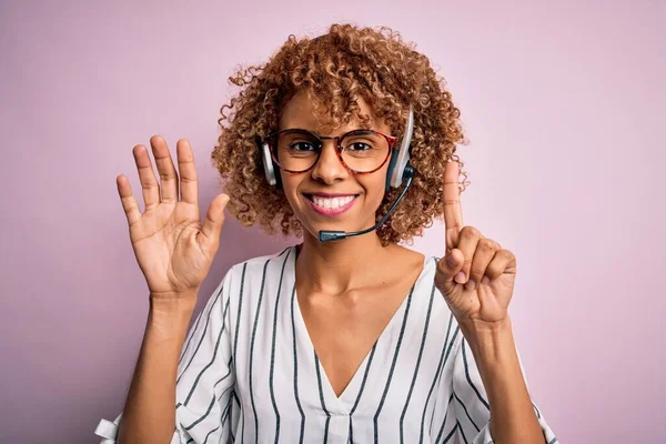 African american curly call center agent woman working using headset over pink background showing and pointing up with fingers number six while smiling confident and happy.