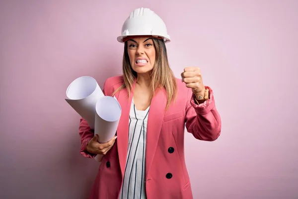 Young beautiful brunette architect woman wearing safety helmet holding blueprints annoyed and frustrated shouting with anger, crazy and yelling with raised hand, anger concept